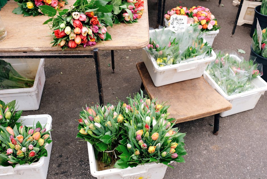 Multiple bouquets of fresh tulips in shades of pink, yellow, white, and red are displayed in white plastic crates and on wooden tables at an outdoor flower market. The flowers have vibrant, opened pet