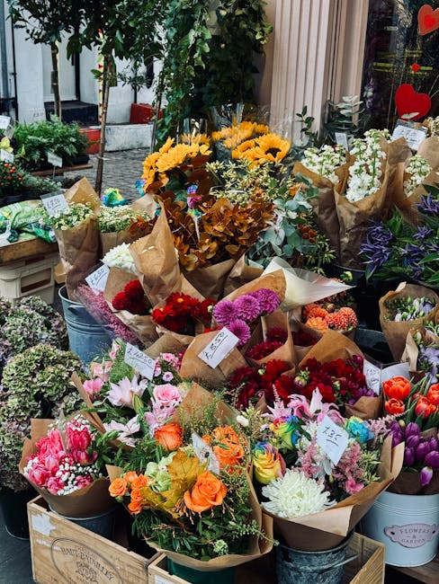 A vibrant display of freshly arranged flower bouquets sit outdoors, featuring a variety of blooms such as bright yellow sunflowers, pink lilies, deep red and purple carnations, orange roses, and white