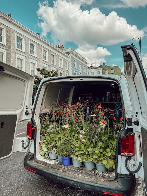 The back of a white delivery van open to reveal a variety of fresh flowers and foliage arranged in blue and gray pots. The flowers include tall, delicate red poppies, white daisies, and lush green lea
