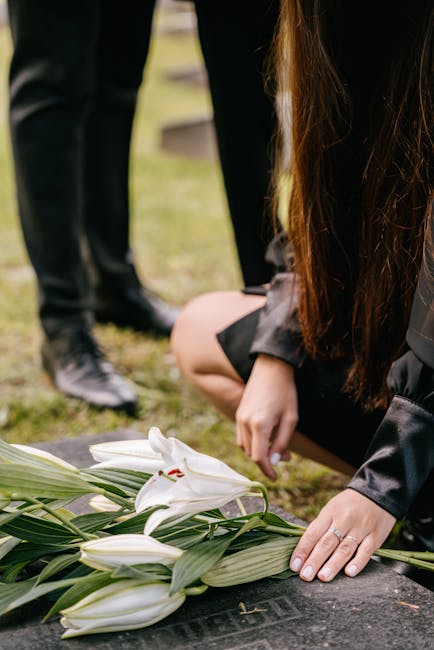 A person with long brown hair wearing a dark jacket is kneeling at a grave, placing a floral arrangement of white lilies with green leaves on a stone marker. The lilies are fresh with open petals, and