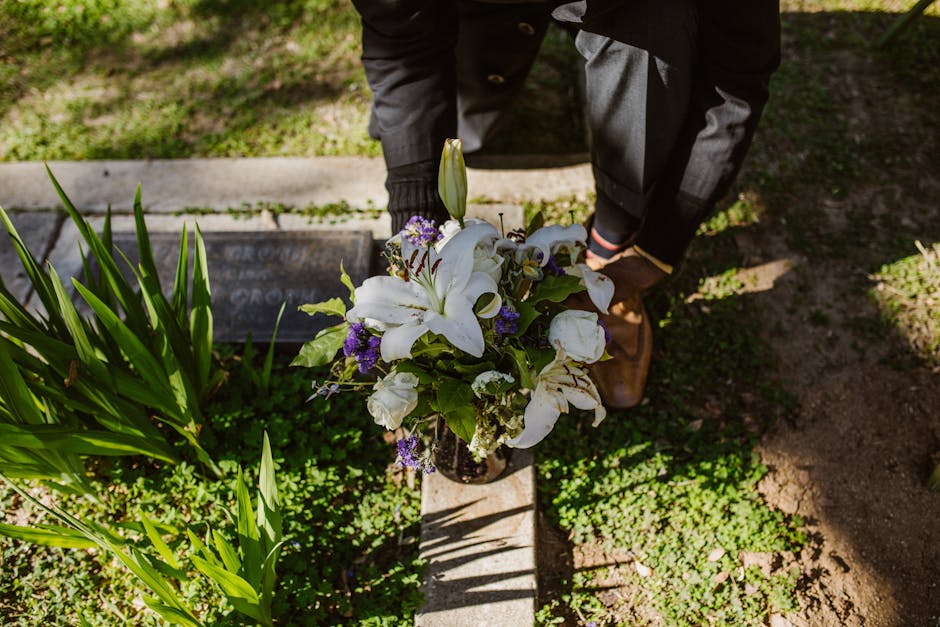 A person dressed in dark clothing, including a coat and trousers, is kneeling on the ground during daylight, arranging a floral bouquet consisting of large white lilies, white roses, and small purple