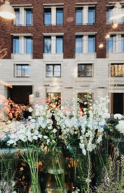 View through a florist shop window showing a variety of fresh floral arrangements, including white and pale pink flowers such as lilies, roses, and snapdragons, set against a backdrop of a red-brick b