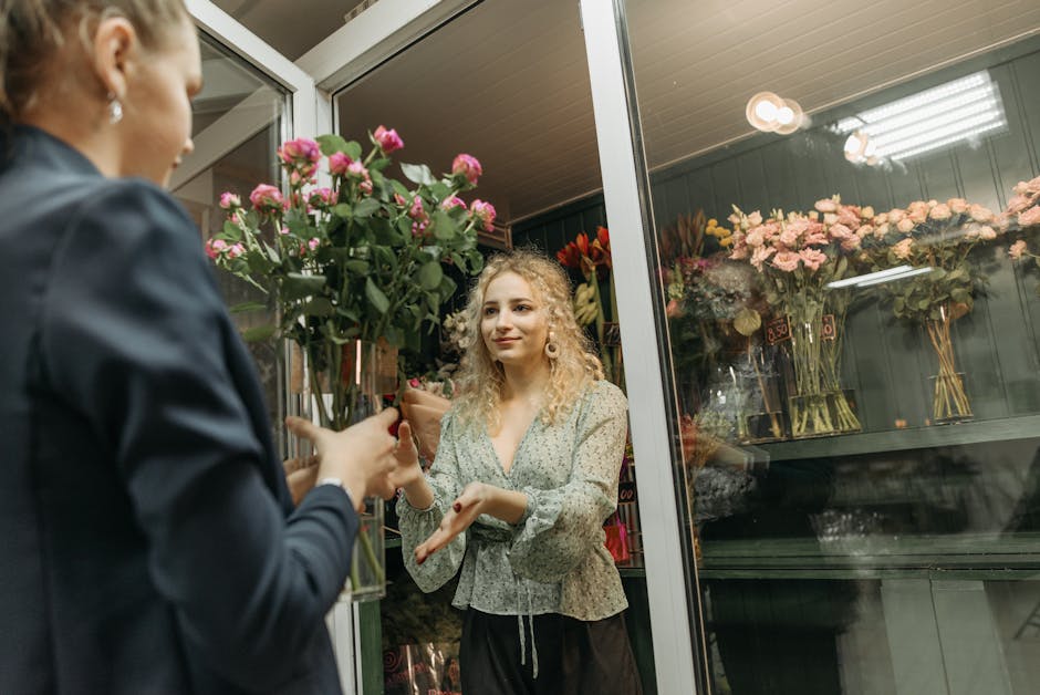 A woman with blonde curly hair wearing a patterned blouse is receiving a bouquet of pink roses from a florist through a glass shop door. The bouquet is wrapped in brown paper with a white ribbon, and 