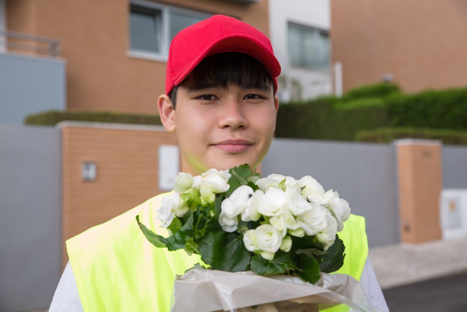 A young delivery person wearing a red cap and a neon yellow vest holding a bouquet of fresh white roses wrapped in white paper. The arrangement features multiple roses with lush green leaves, showcasi